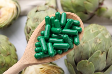 Photo of Wooden spoon of pills over table with fresh artichokes, closeup