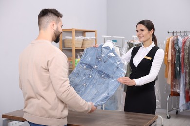 Photo of Dry-cleaning service. Happy worker giving denim jacket in plastic bag to client indoors