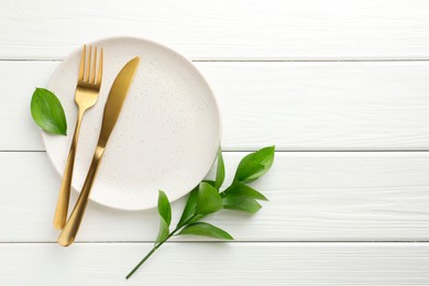 Stylish setting with cutlery, green leaves and plate on white wooden table, flat lay. Space for text