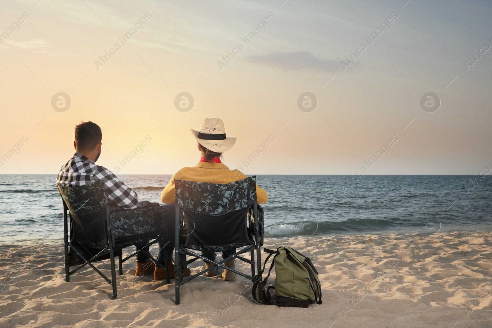 Photo of Couple sitting in camping chairs and enjoying seascape on beach, back view