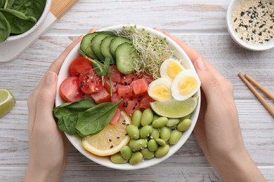 Woman holding delicious poke bowl with quail eggs, fish and edamame beans at white wooden table, top view