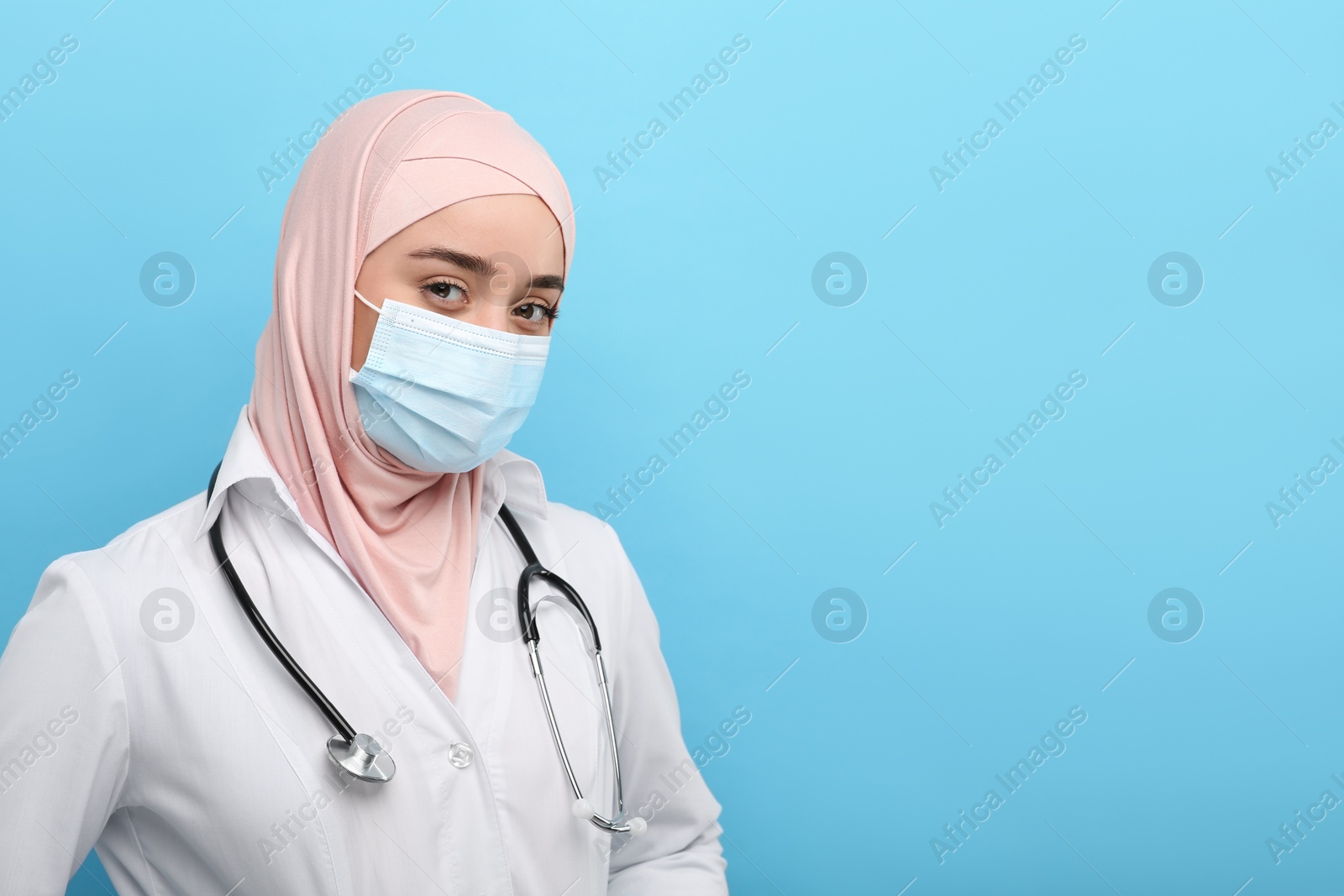 Photo of Muslim woman wearing hijab, medical uniform and protective mask on light blue background, space for text