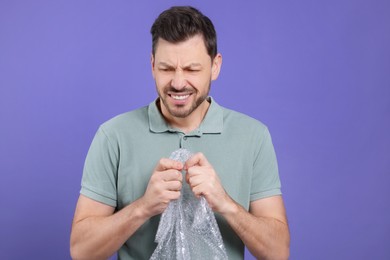 Angry man popping bubble wrap on purple background. Stress relief