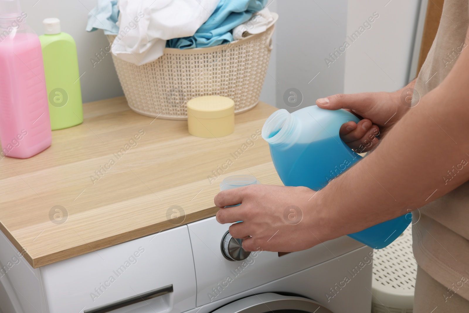 Photo of Man pouring fabric softener from bottle into cap near washing machine indoors, closeup