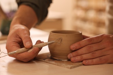 Photo of Clay crafting. Man making bowl with sculpting tool at table in workshop, closeup