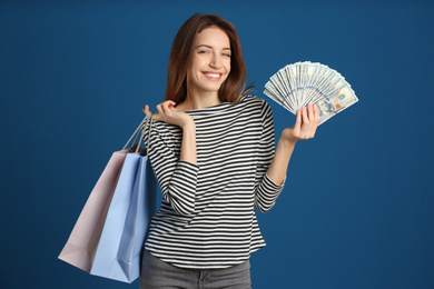 Happy young woman with cash money and shopping bags on blue background