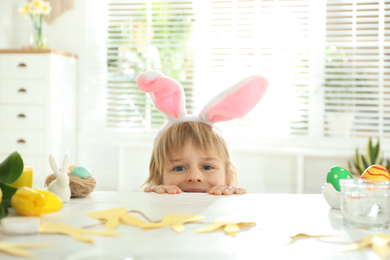 Photo of Cute little boy wearing bunny ears headband at table with Easter eggs, indoors