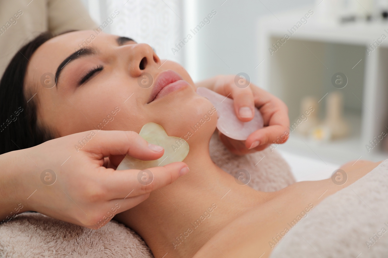 Photo of Young woman receiving facial massage with gua sha tools in beauty salon