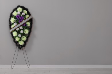 Funeral wreath of plastic flowers near grey wall indoors. Space for text