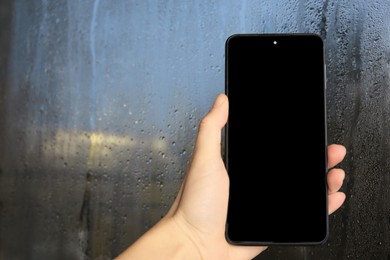 Photo of Woman holding smartphone near window with rain drops, closeup. Space for text