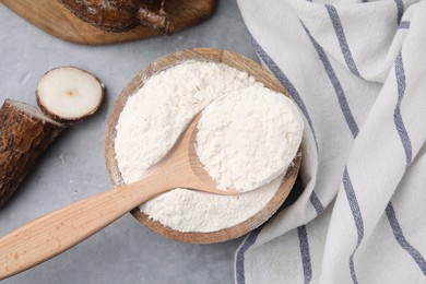 Wooden bowl, spoon with cassava flour and roots on grey table, flat lay