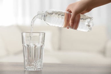 Woman pouring water from bottle into glass at table on blurred background, closeup
