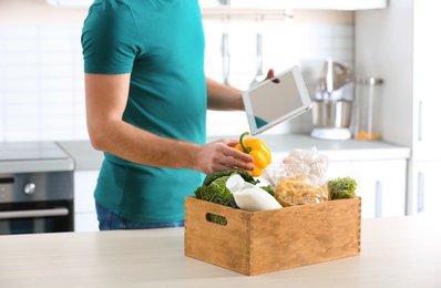 Man with wooden crate full of products and tablet in kitchen. Food delivery service