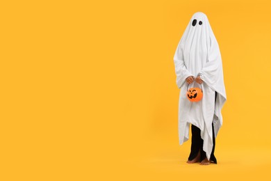 Photo of Woman in white ghost costume holding pumpkin bucket on yellow background, space for text. Halloween celebration