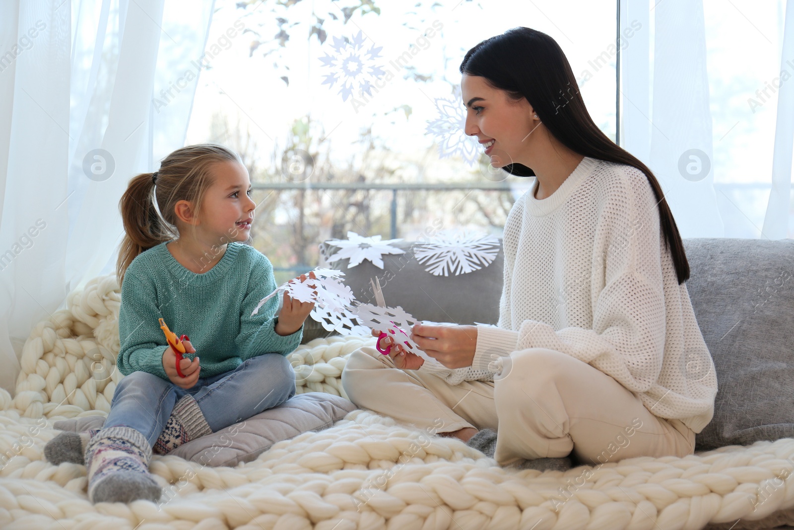 Photo of Mother and daughter making paper snowflakes near window at home