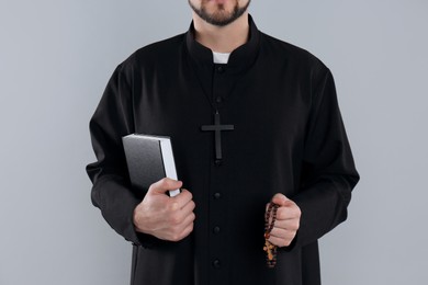 Priest with Bible and rosary beads on grey background, closeup