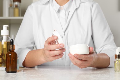 Photo of Dermatologist holding jar of cream at white table indoors, closeup. Developing cosmetic product