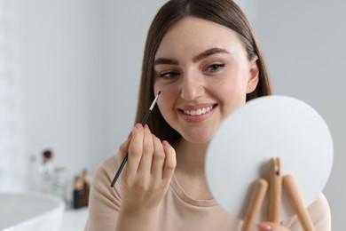 Photo of Smiling woman drawing freckles with brush in front of little mirror indoors