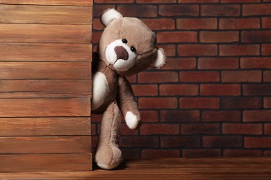 Photo of Cute teddy bear peeking out behind wooden wall against brick background, space for text