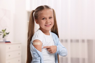 Photo of Happy girl pointing at sticking plaster after vaccination on her arm indoors