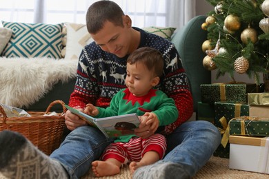 Photo of Father reading book to his cute son in room decorated for Christmas