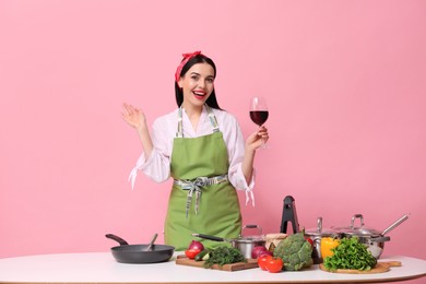 Photo of Young housewife with glass of wine, vegetables and different utensils on pink background