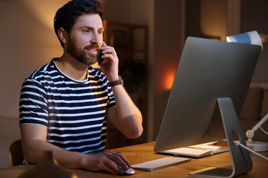 Photo of Home workplace. Man talking on smartphone while working with computer at wooden desk at night