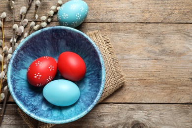 Photo of Flat lay composition of bowl with painted Easter eggs and pussy willow on wooden table, space for text