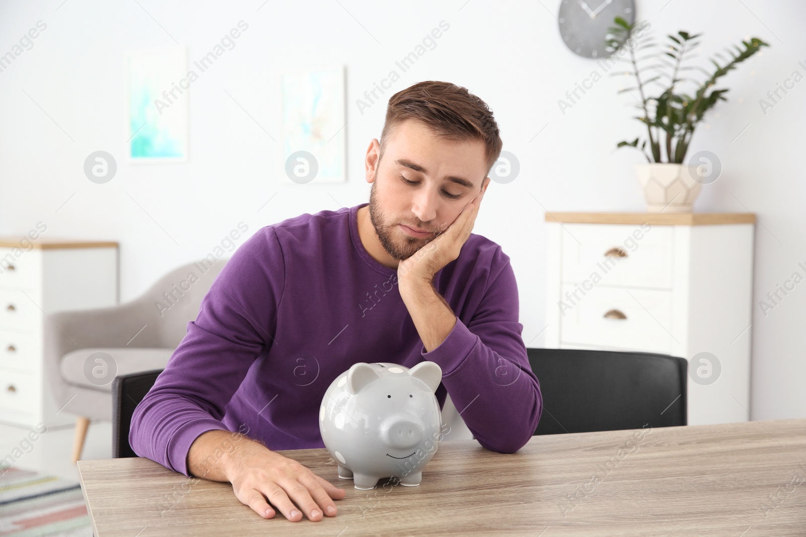 Photo of Unhappy young man with piggy bank at table indoors