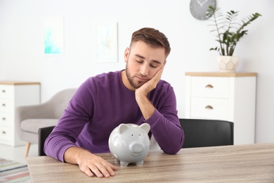 Photo of Unhappy young man with piggy bank at table indoors