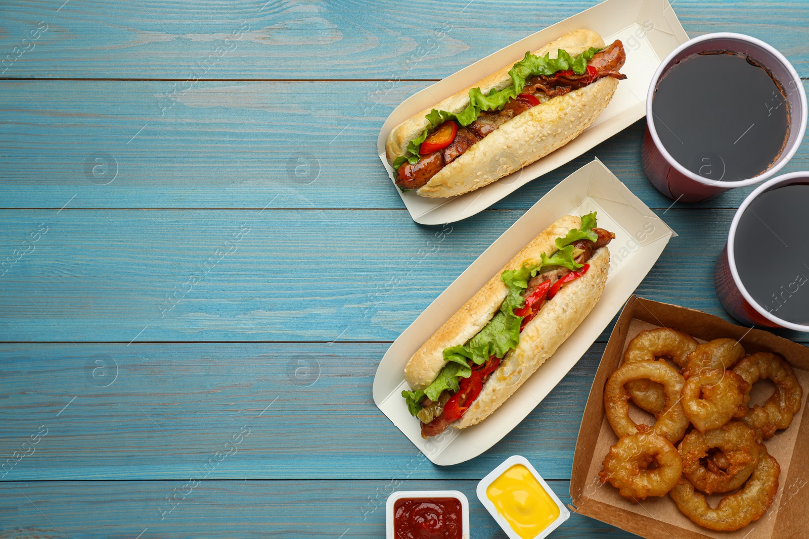 Photo of Tasty hot dogs, fried onion rings, sauces and refreshing drink on light blue wooden table, flat lay with space for text. Fast food