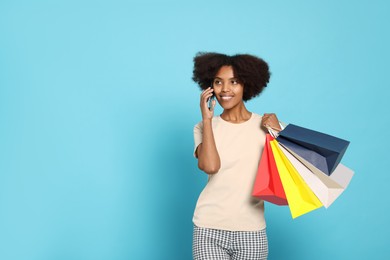 Happy African American woman with shopping bags talking on smartphone against light blue background. Space for text