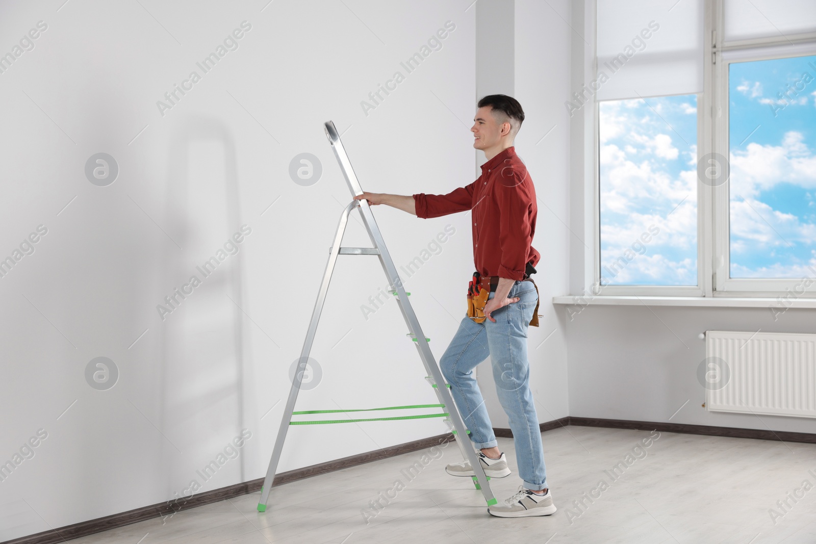 Photo of Young handyman with stepladder and tool belt working in room