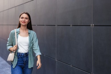 Young woman in casual clothes walking near grey wall outdoors, space for text