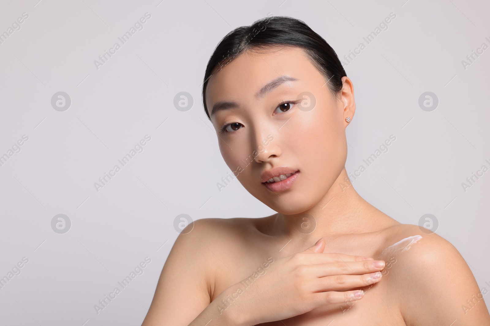 Photo of Beautiful young Asian woman applying body cream onto shoulder on light grey background, space for text