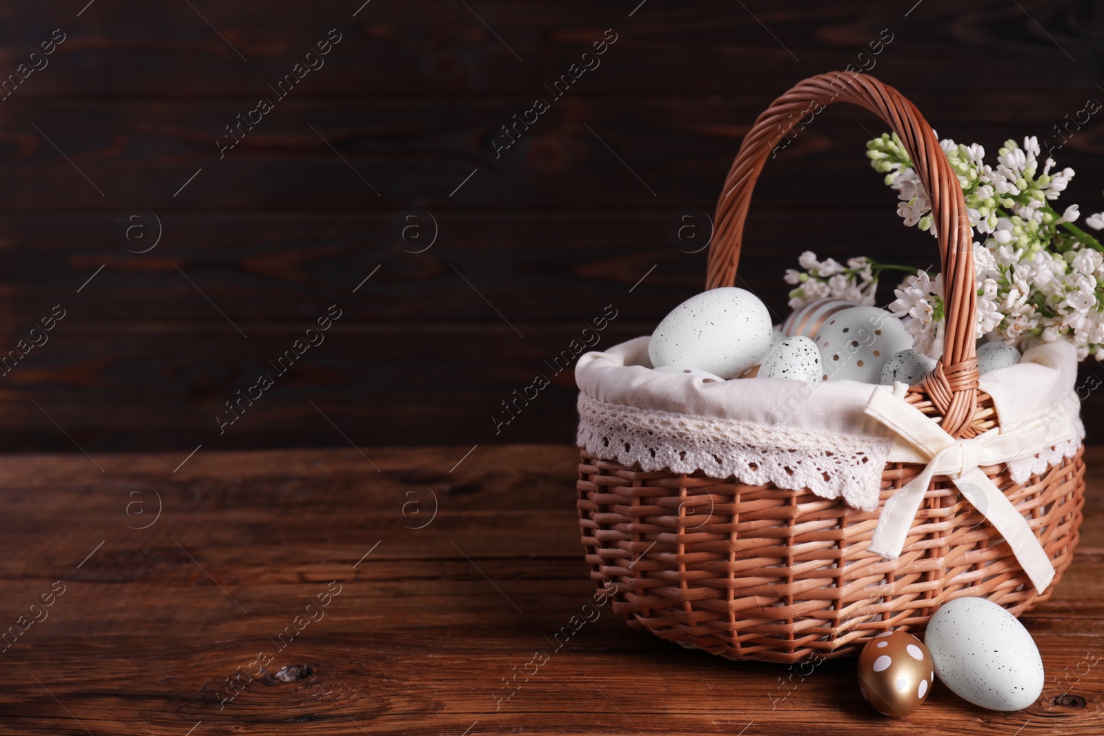 Photo of Wicker basket with festively decorated Easter eggs and white lilac flowers on wooden table. Space for text