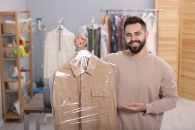 Photo of Dry-cleaning service. Happy man holding hanger with jacket in plastic bag indoors