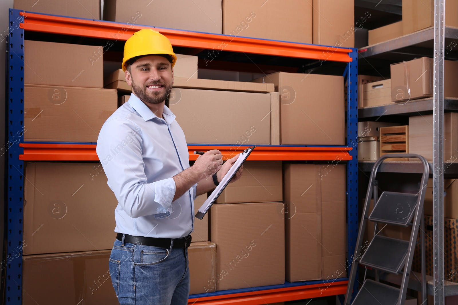 Photo of Young man with clipboard near rack of cardboard boxes at warehouse