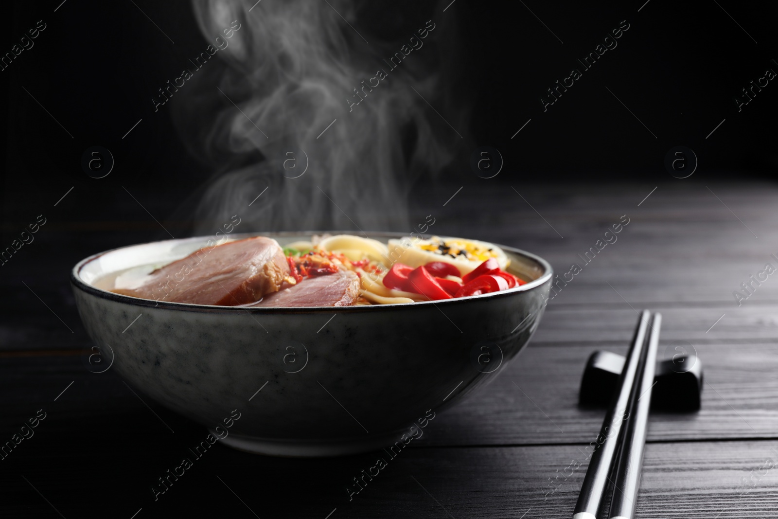 Image of Delicious hot ramen in bowl and chopsticks on black wooden table, closeup. Noodle soup