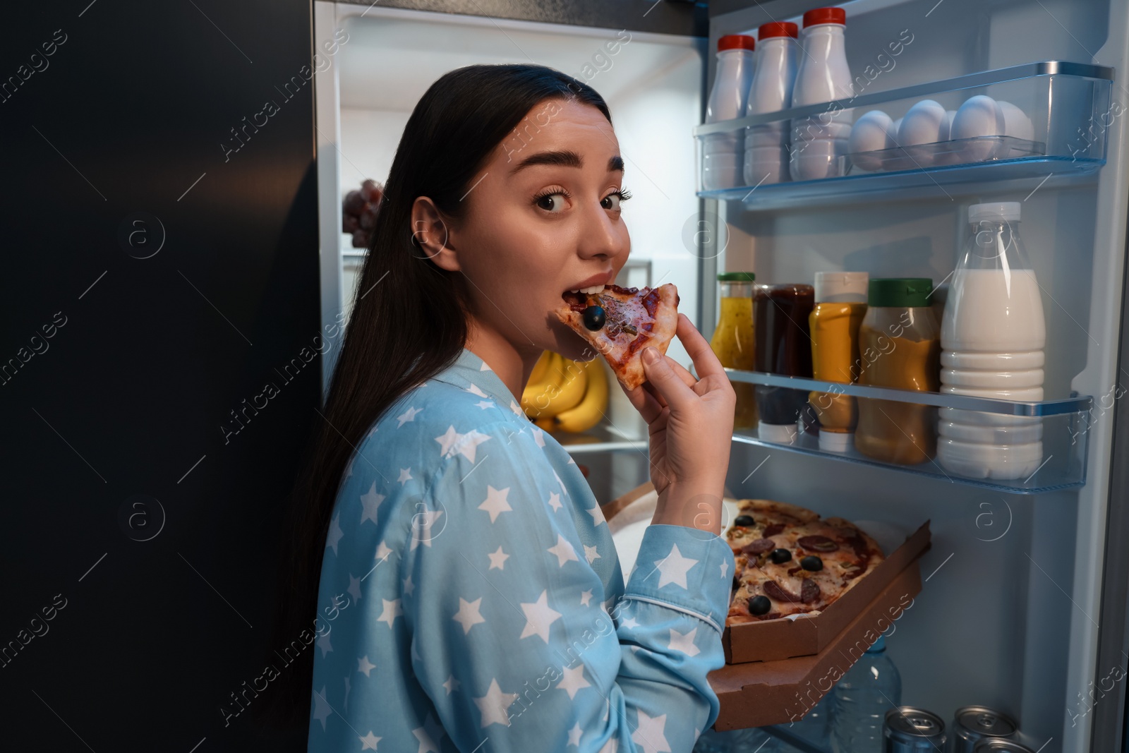 Photo of Young woman eating pizza near refrigerator in kitchen at night. Bad habit