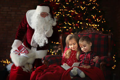 Photo of Santa Claus sneaking in with gifts while children reading book near Christmas tree indoors