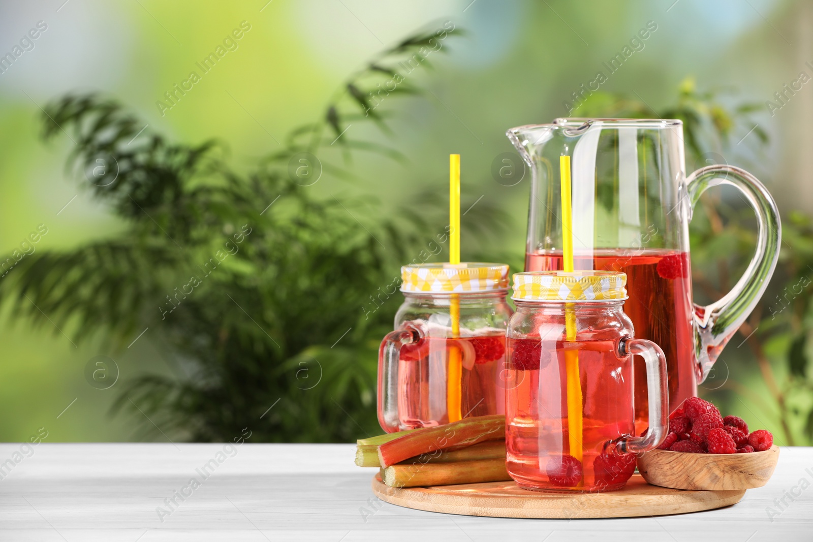 Photo of Tasty rhubarb cocktail with raspberry and stalks on white wooden table outdoors, space for text