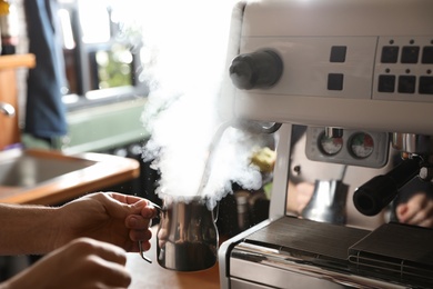 Photo of Barista steaming milk in metal jug with coffee machine wand at bar counter, closeup