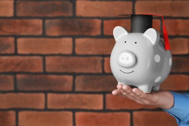 Photo of Man holding piggy bank and graduation cap against brick background, closeup with space for text. Scholarship concept