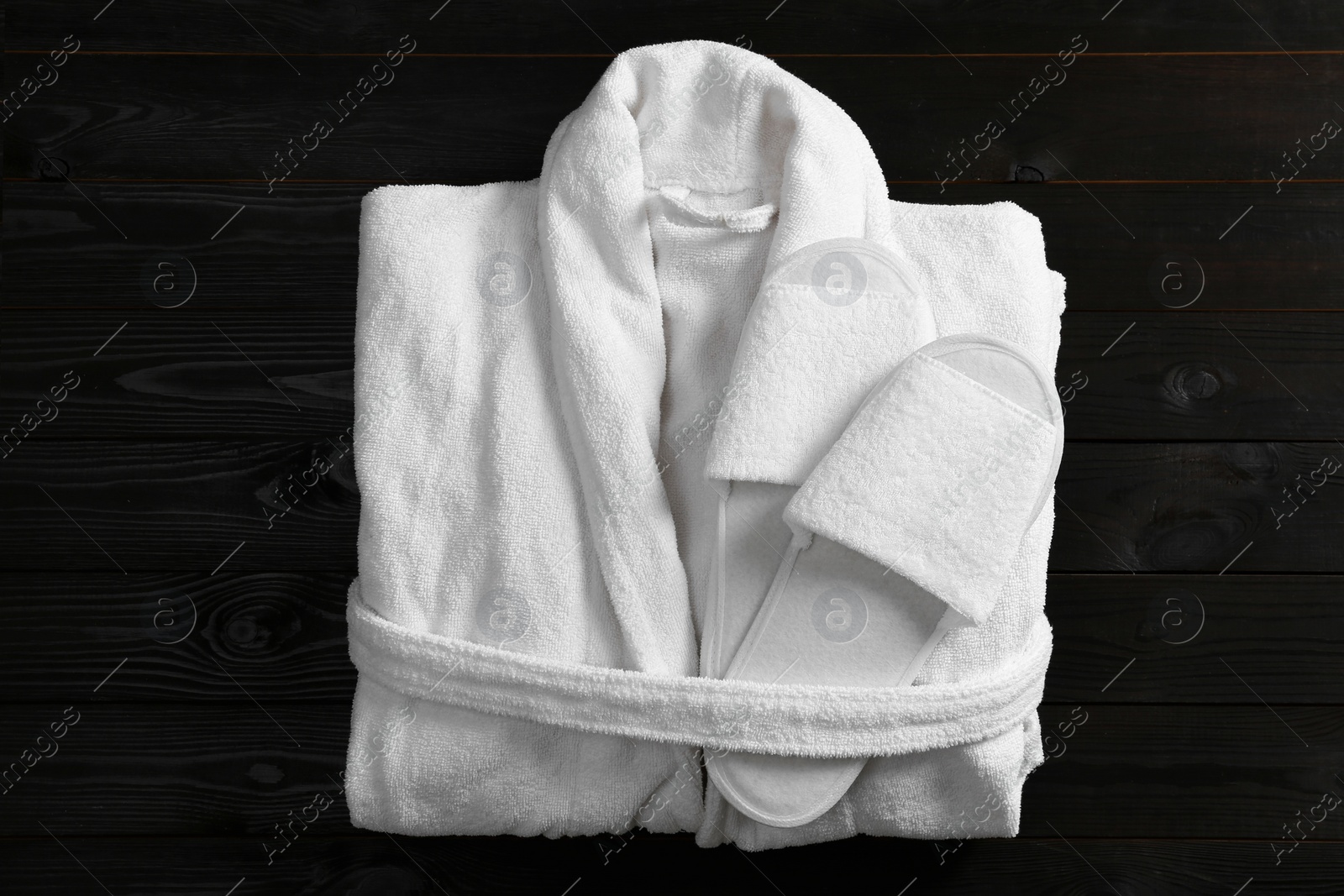 Photo of Clean folded bathrobe and slippers on black wooden background, top view