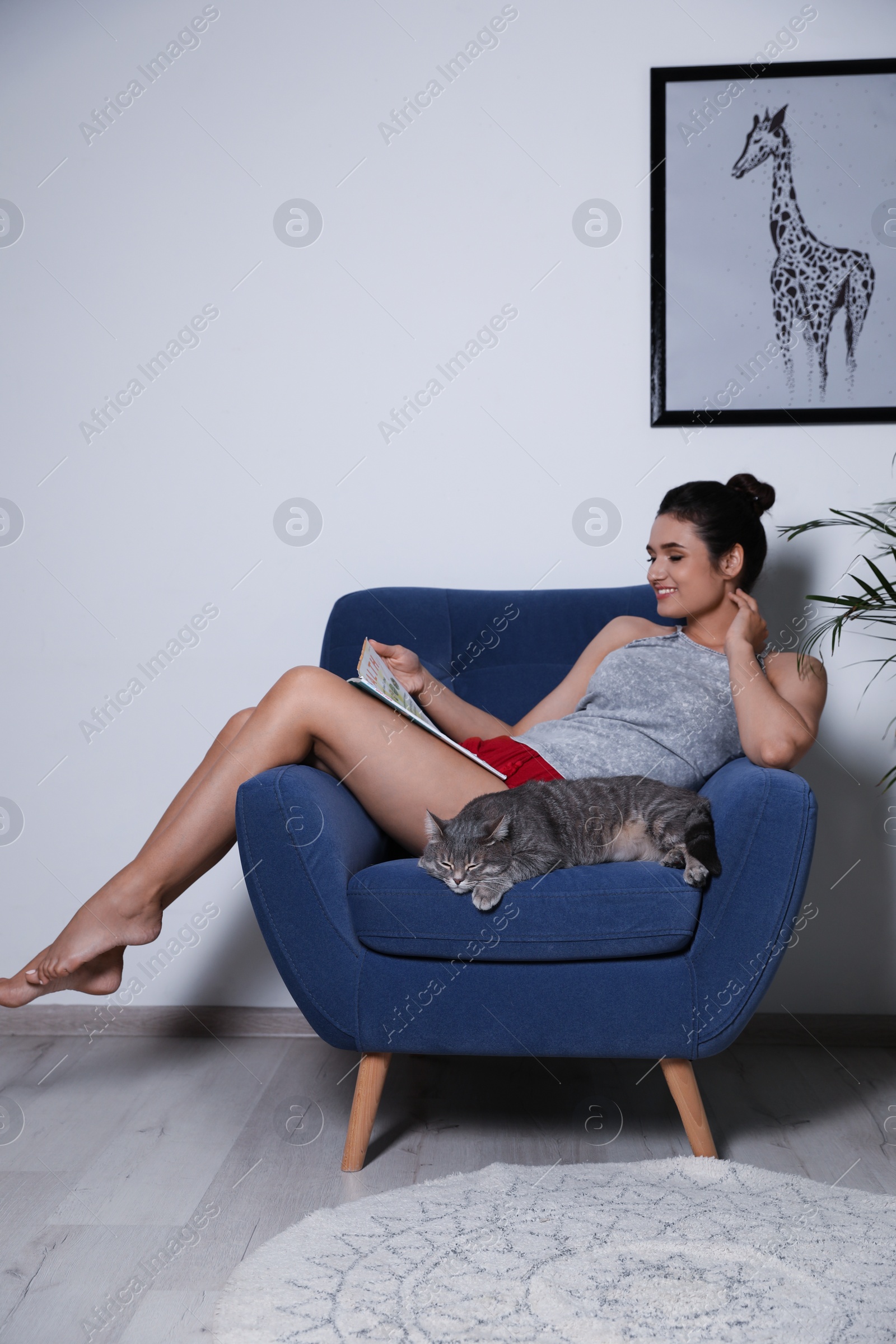 Photo of Young woman with cat and magazine on armchair at home. Cute pet