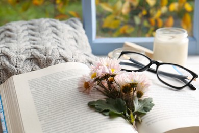 Photo of Open book with chamomile flowers as bookmark, scented candle and glasses on table near window, closeup