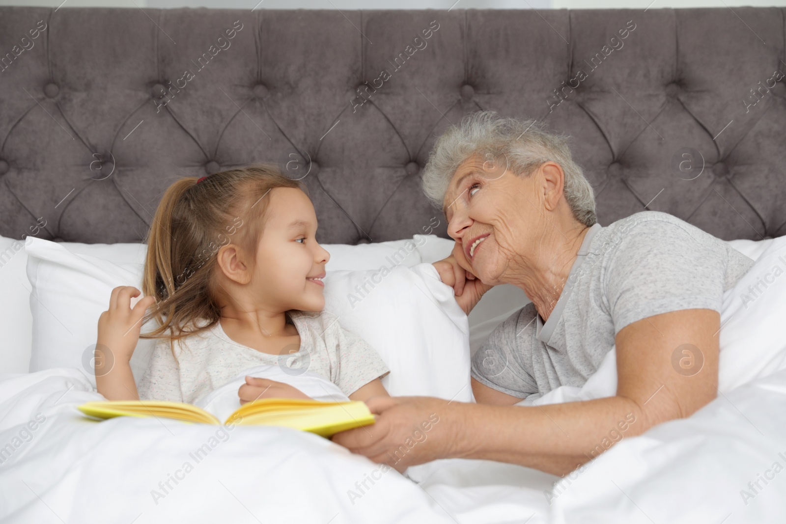 Photo of Cute girl and her grandmother reading book on bed at home