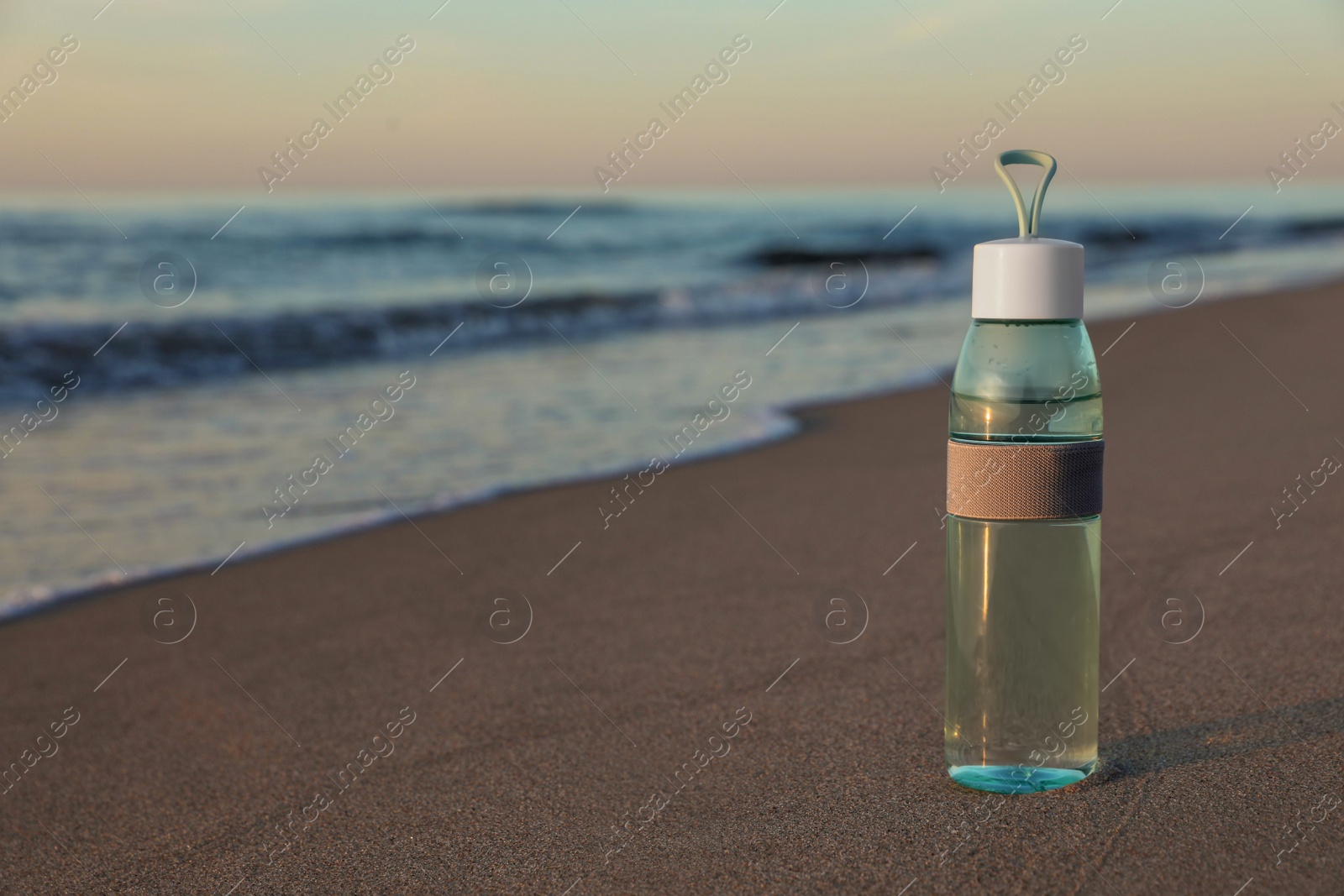 Photo of Glass bottle with water on wet sand near sea at sunset. Space for text