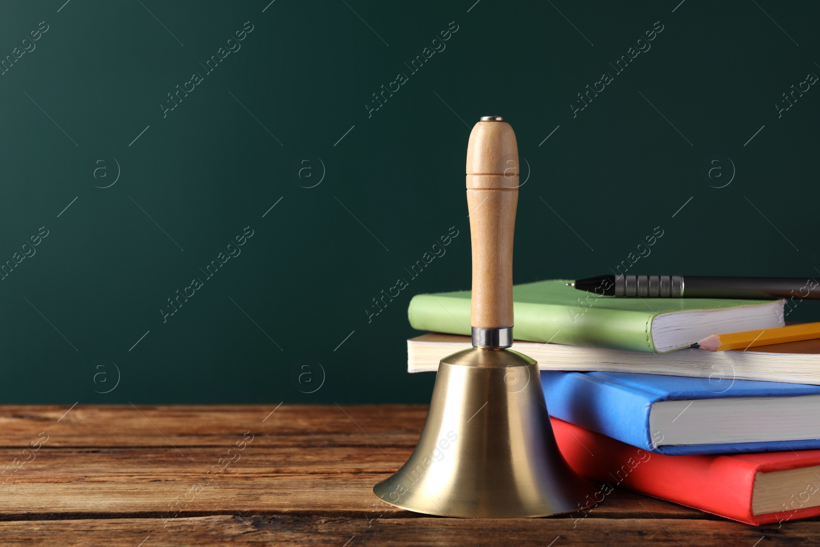Photo of Golden bell, books and stationery on wooden table near green chalkboard, space for text. School days